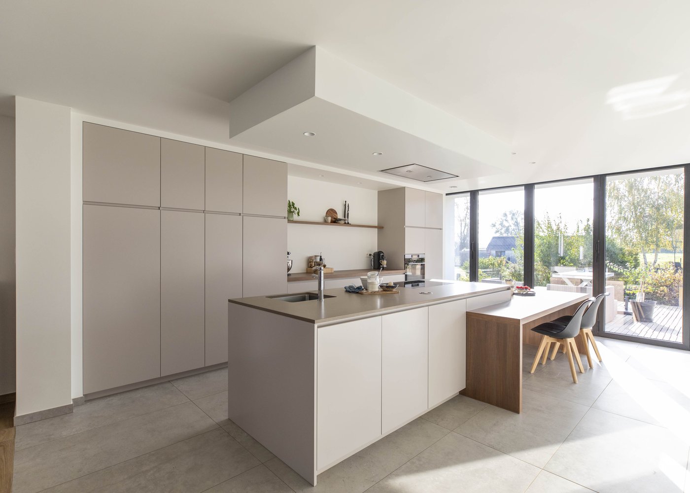Op de foto zie je een moderne, strak vormgegeven leefkeuken met een centraal kookeiland en een houten verlengstuk dat naadloos overloopt in de eettafel. Het gaat om een maatwerk keukenservice die interieurontwerp en renovatie combineert. Het hoofddoel van deze dienst is het realiseren van een functionele en stijlvolle keuken die naadloos aansluit op de rest van de woonruimte. De greeploze kastdeuren zorgen voor een minimalistische uitstraling en extra bergplaats, de geïntegreerde spoelbak in het werkblad maakt schoonmaken eenvoudiger en de grote glazen schuifdeuren bieden maximale lichtinval en verbinden de keuken direct met de tuin.